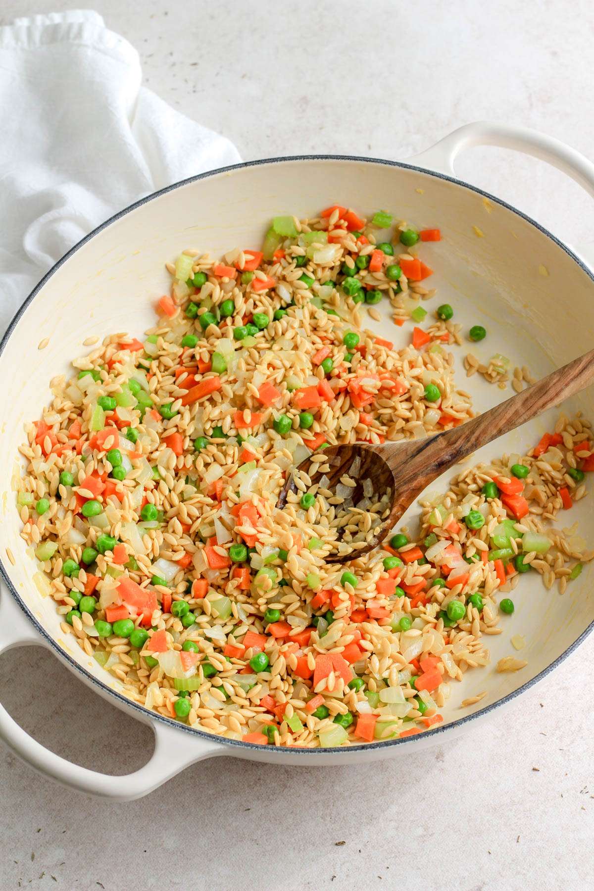A white pan with a wooden spoon and cooked vegetables with uncooked orzo.