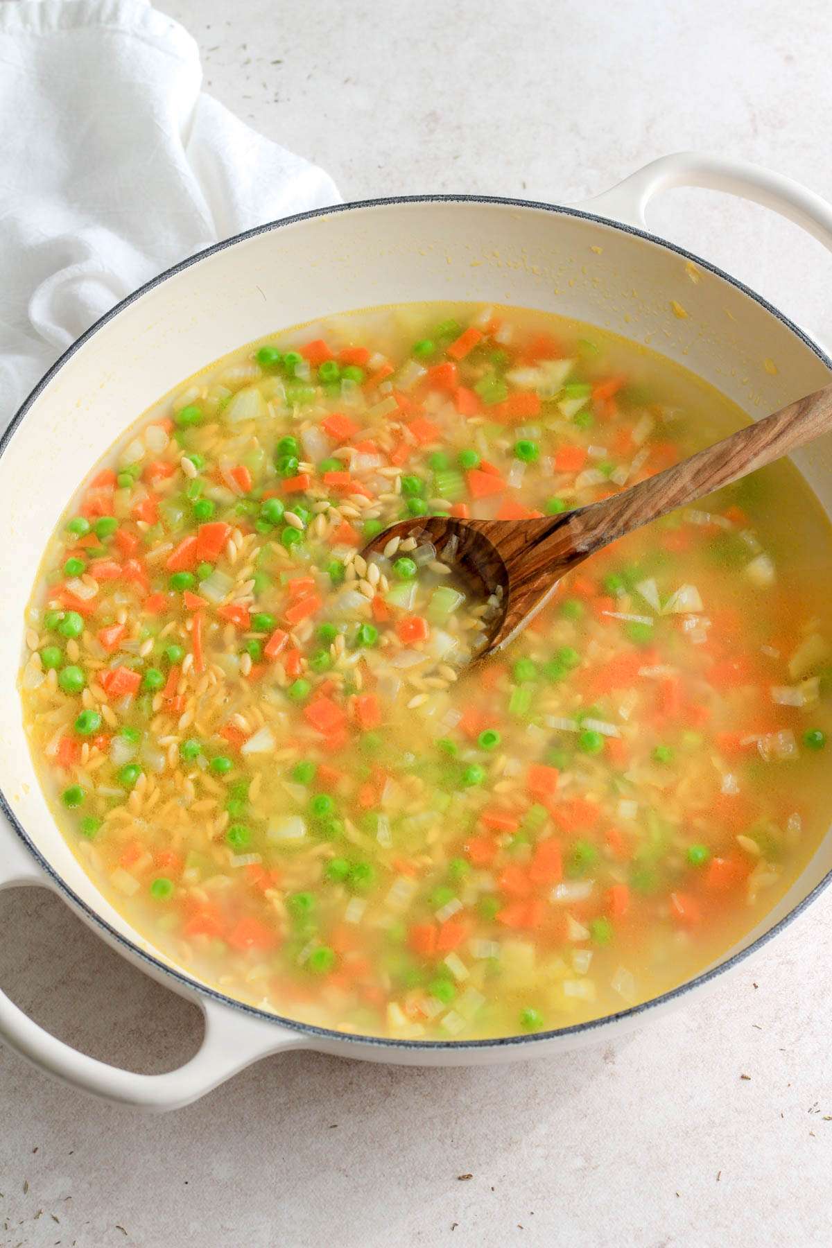 A white pan with a wooden spoon and the vegetables with uncooked orzo and chicken broth.