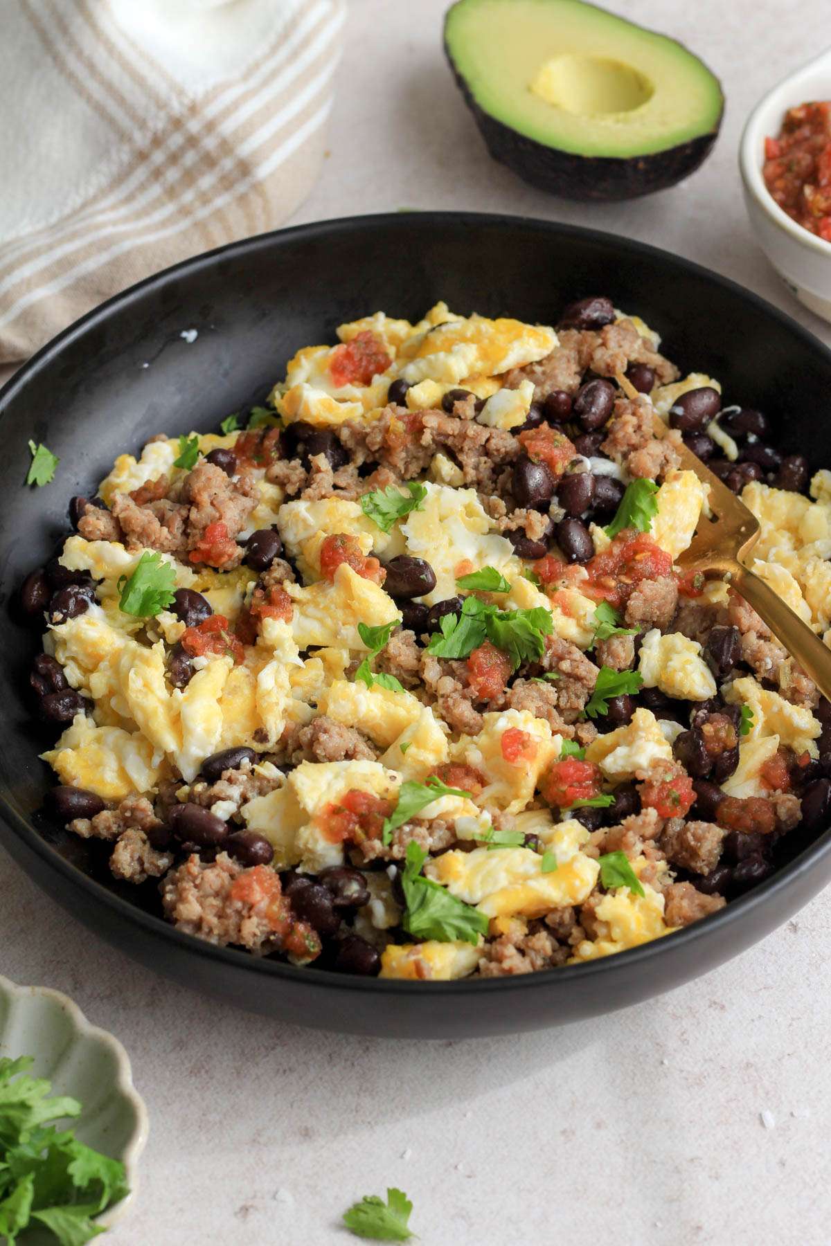 From the side angle of a black bowl with the sausage, black bean and scrambled egg bowl topped with cilantro and salsa on a tan counter.