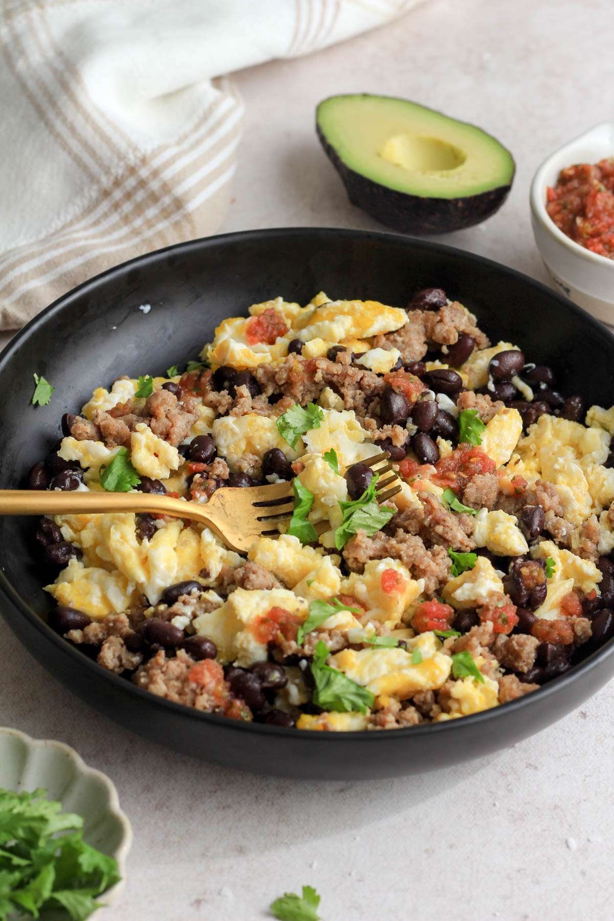 A gold fork in a black bowl of black bean and egg scramble on a tan counter with avocado, cilantro, and salsa around the bowl.