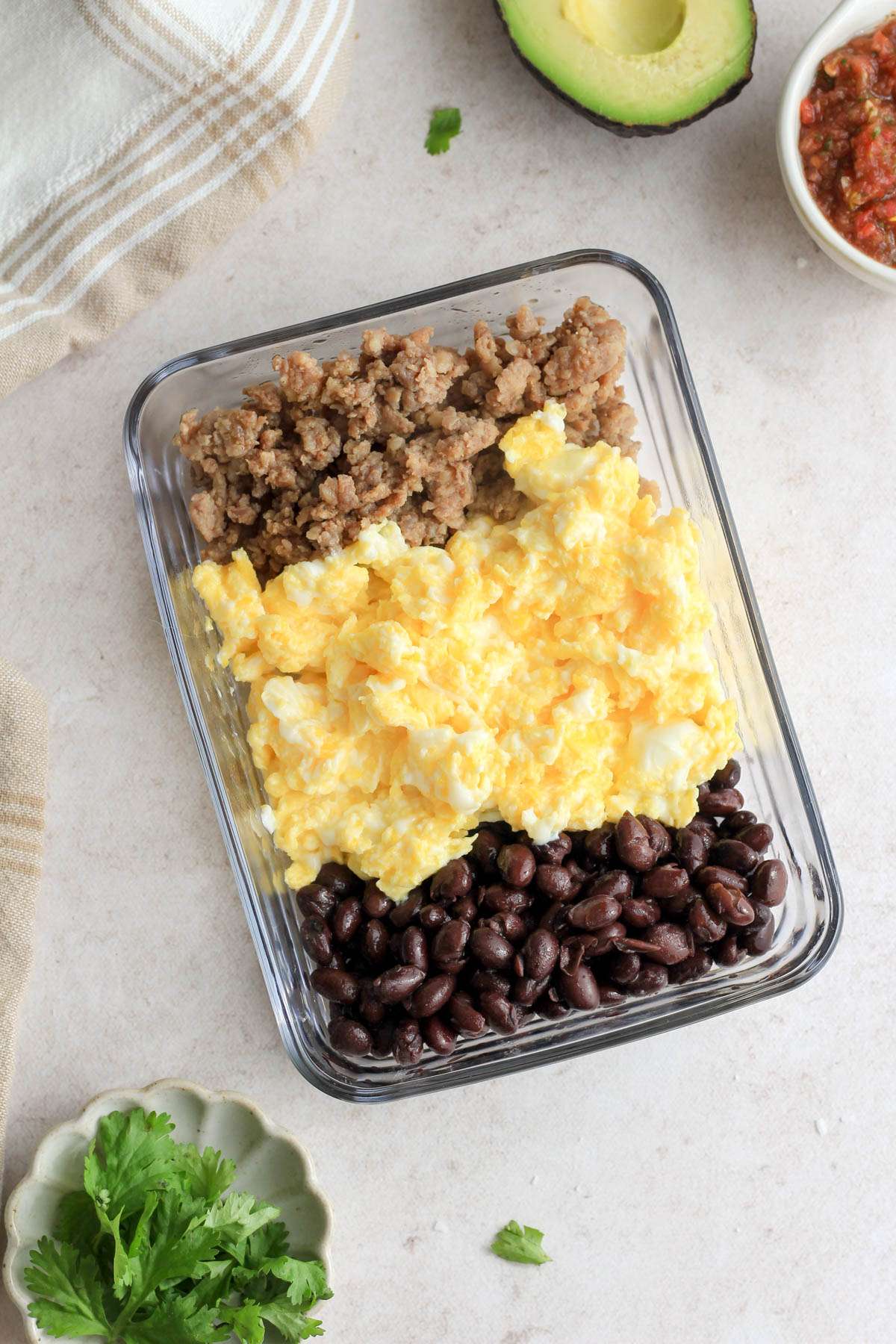 A top down image of a meal prep container with breakfast sausage, scrambled eggs, and black beans on a tan counter with cilantro in the bottom left and salsa and avocado in the top right.