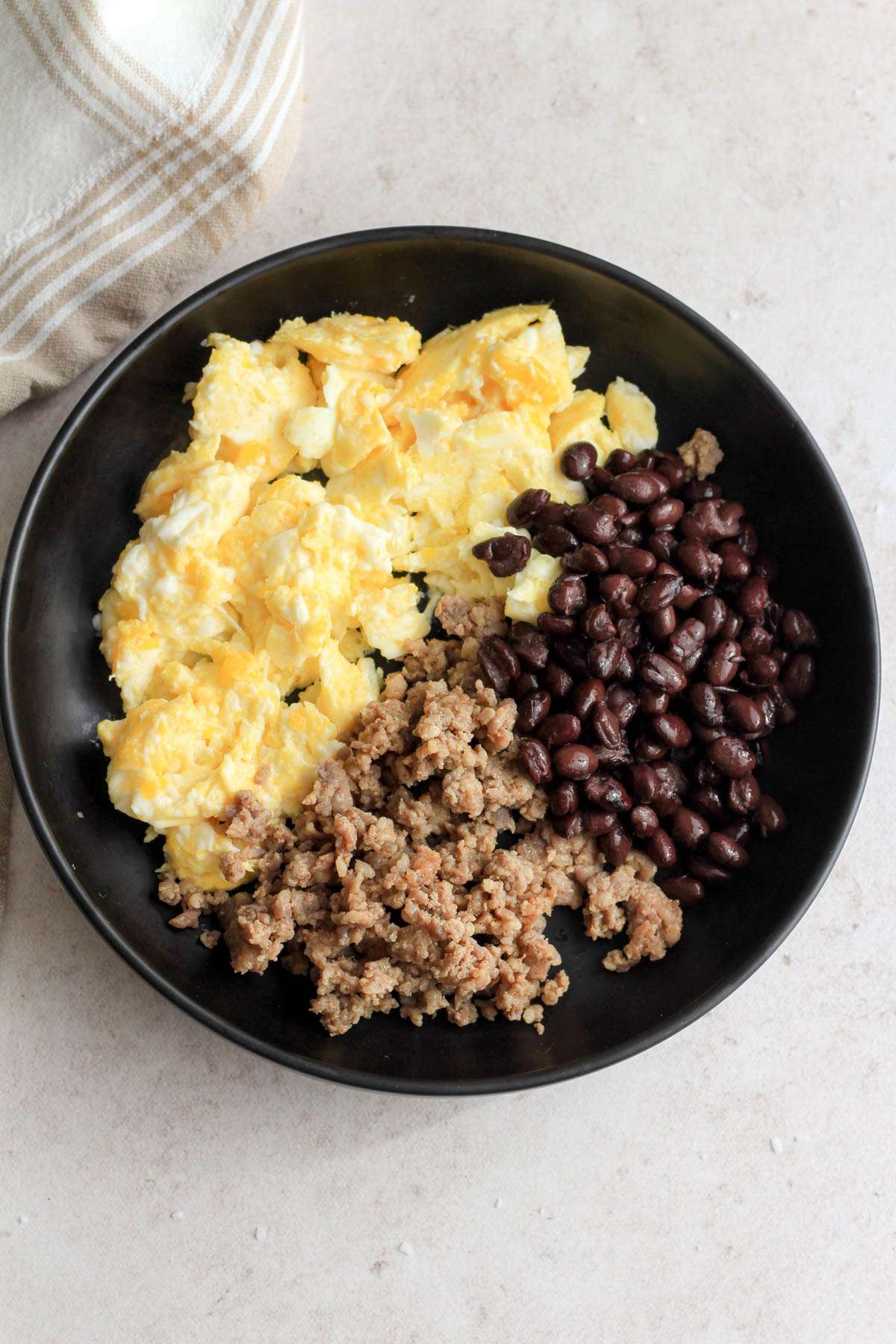 A black bowl with scrambled eggs, breakfast sausage, and black beans on a tan counter.
