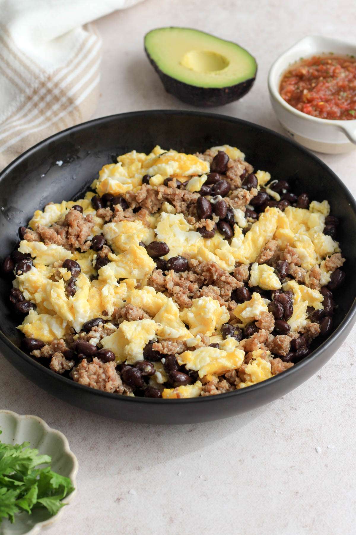 A black bowl with sausage and egg scramble on a tan counter with cilantro, avocado, and salsa.