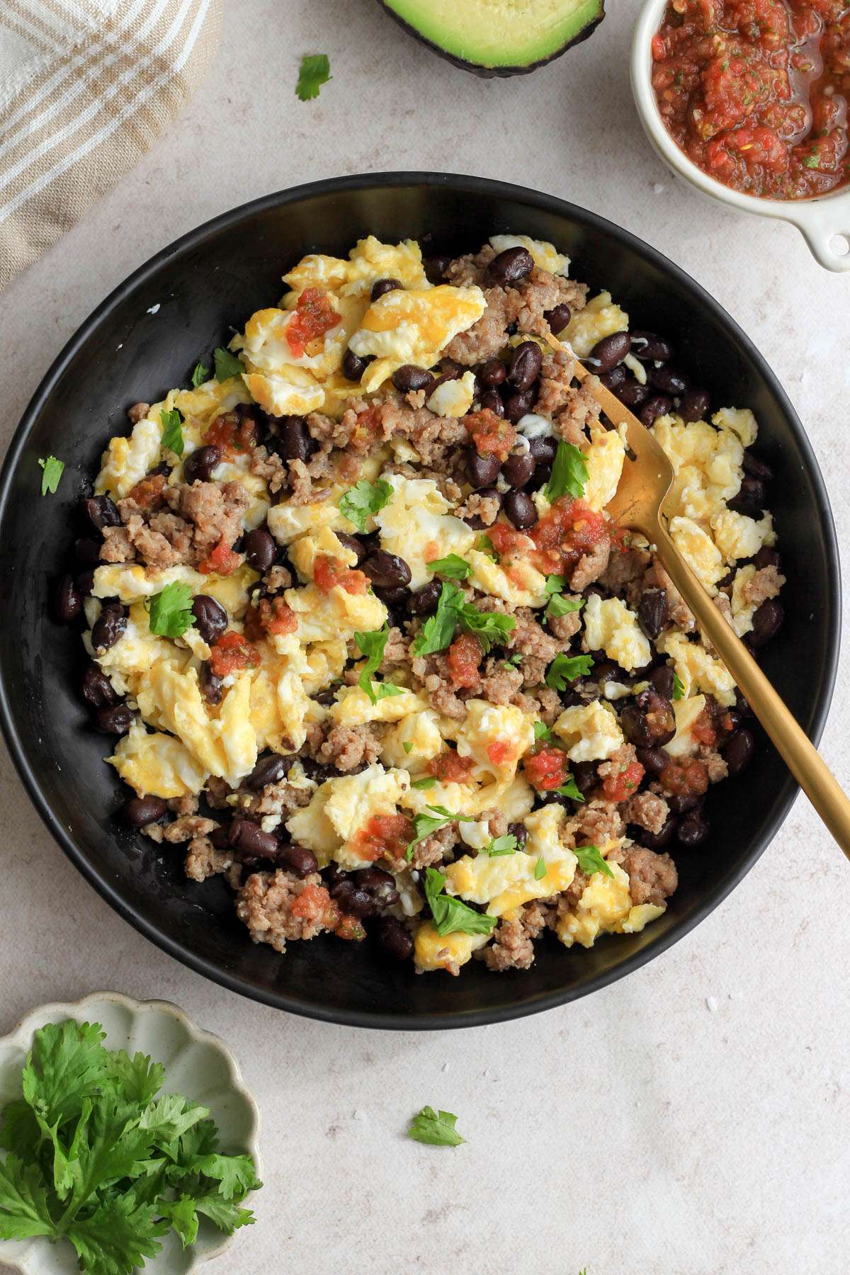 Sausage and scrambled eggs with black beans, cilantro, and salsa in a black bowl with a dish of cilantro in the bottom left and a bowl of salsa in the top right.