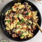 A black bowl with egg and black bean scramble with sausage, cilantro, and salsa with a gold fork in the right.