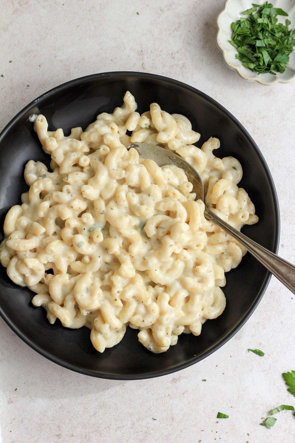 A black bowl filled with vegan Boursin pasta with a silver spoon and a pinch bowl of chopped parsley.