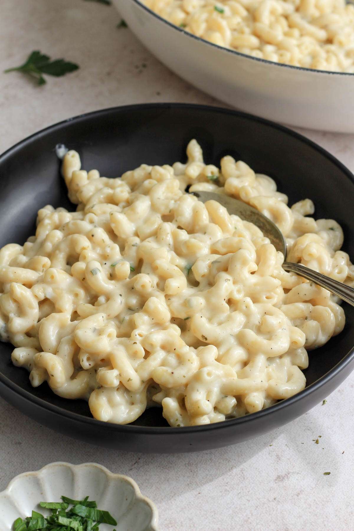 A black bowl of vegan boursin pasta with a silver spoon in front of a white pan with more pasta.