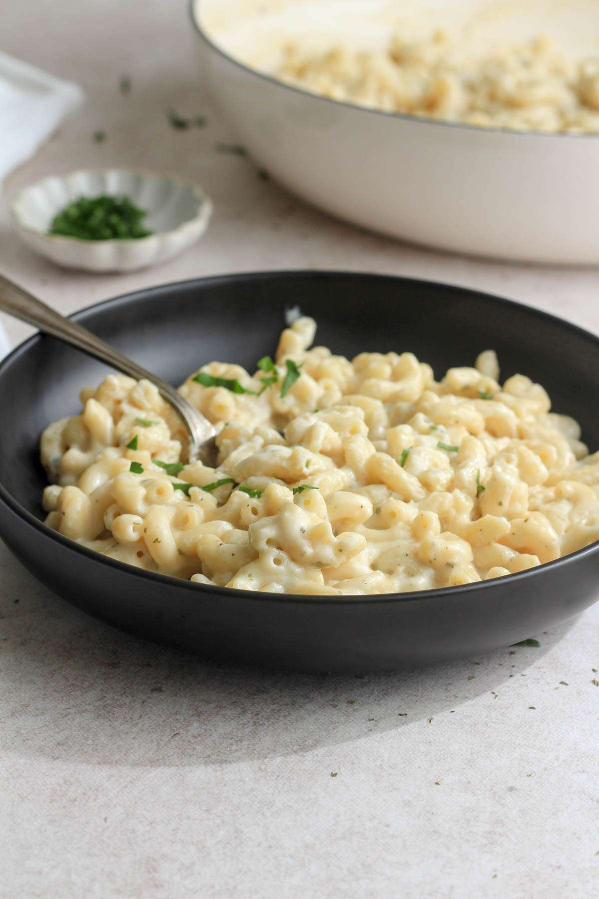 From the side, a black bowl with dairy-free boursin pasta with a spoon on the left side and a white skillet of pasta in the back right.