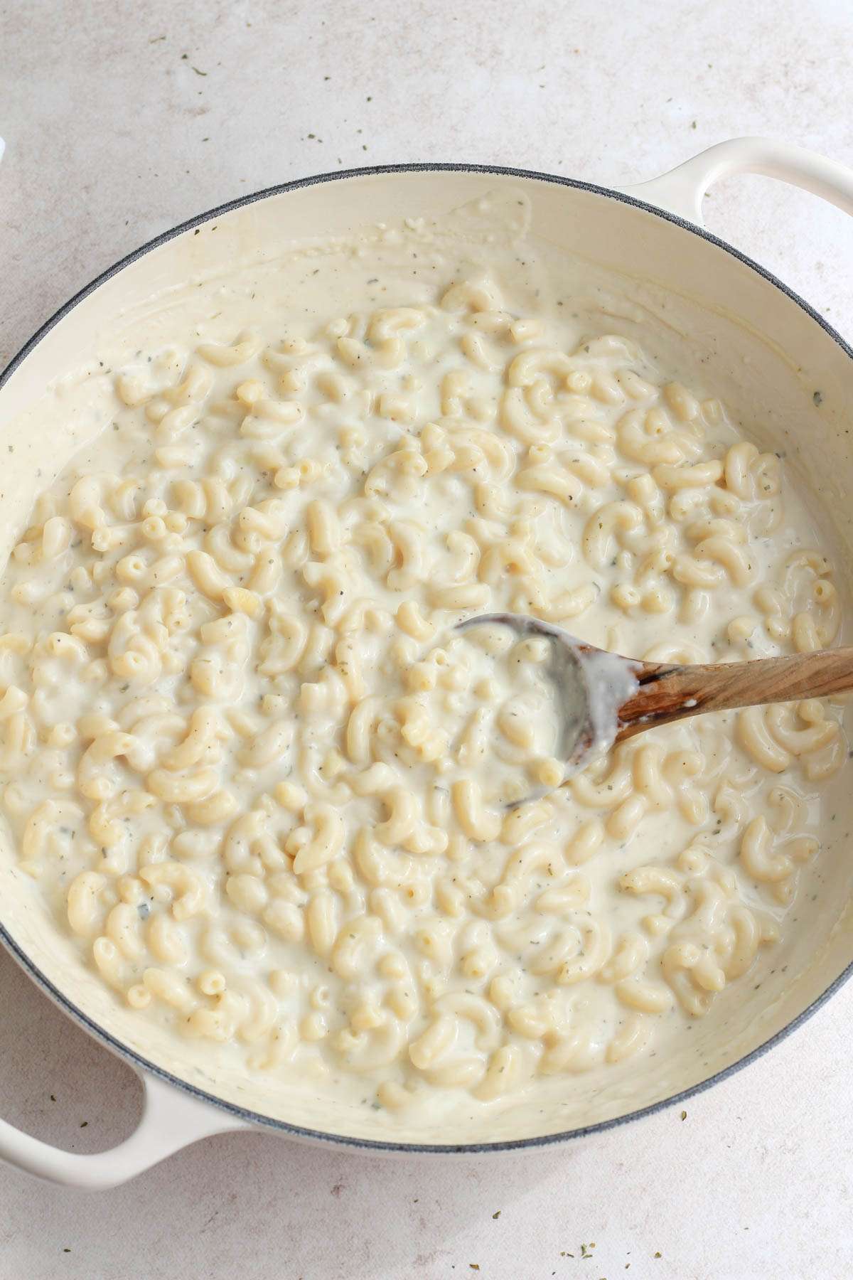 A white skillet with a wooden spoon after adding cooked pasta to the vegan boursin sauce.