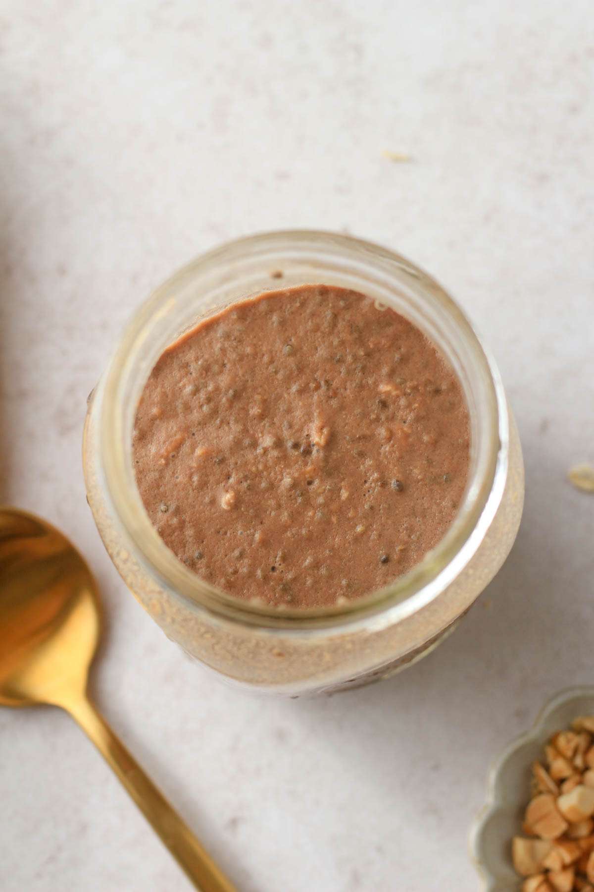 A top down image of chocolate protein powder peanut butter overnight oats in a glass mason jar with a gold spoon to the bottom left.