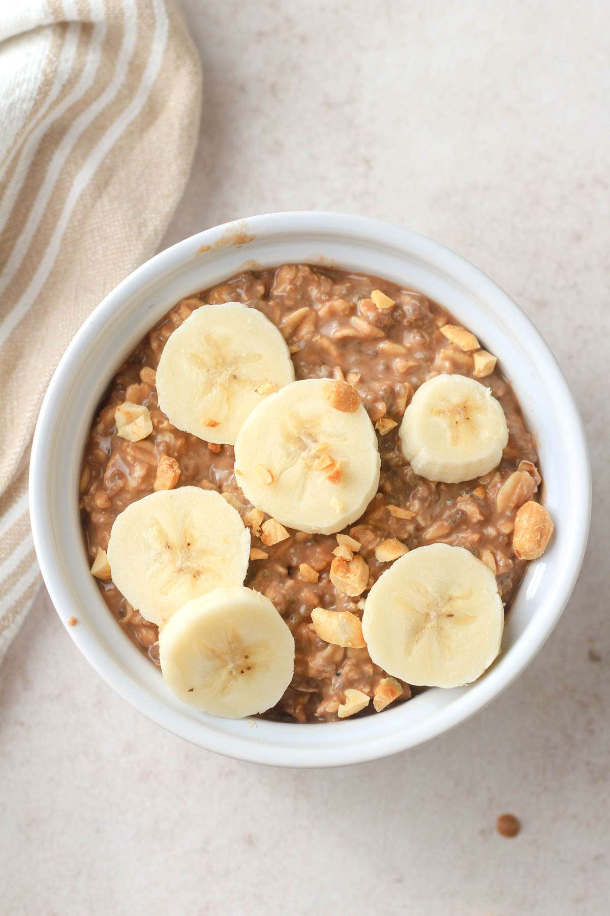 A light tan counter with a white bowl of chocolate peanut butter overnight oats topped with sliced banana and chopped peanuts.