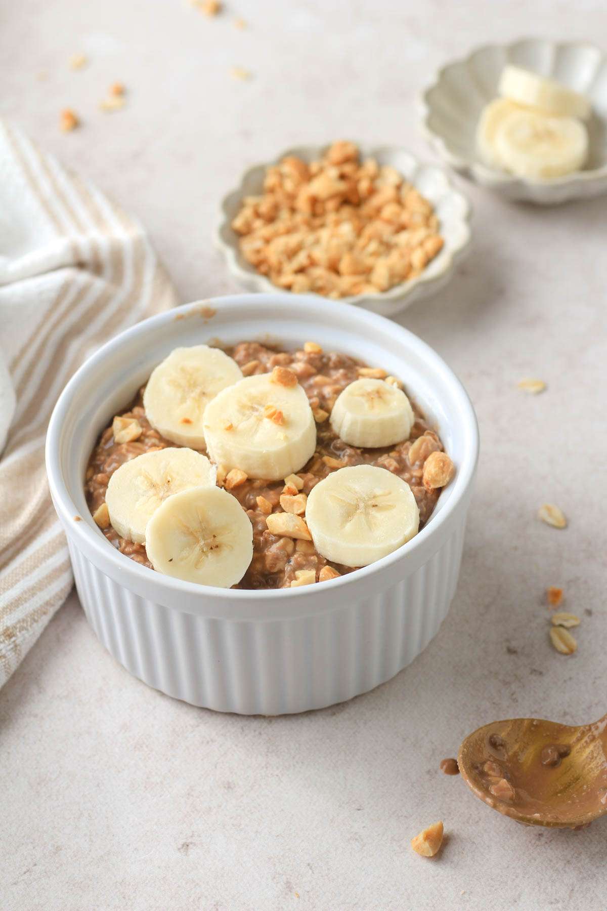 From the side, a white bowl with chocolate protein powder overnight oats topped with banana and a small dish of peanuts and banana in the back.