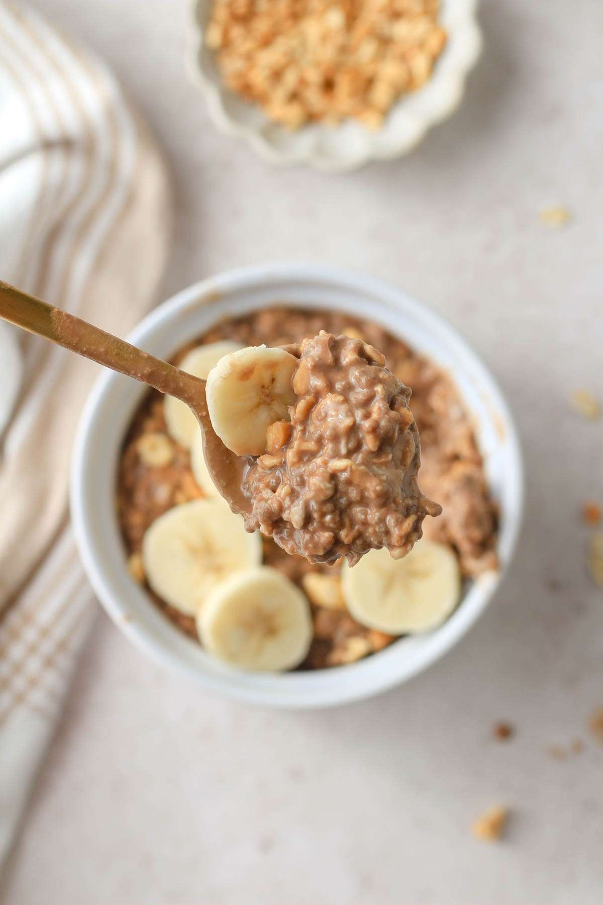 A gold spoon with a scoop of chocolate peanut butter overnight oats and a small slice of banana lifted above a white bowl with dairy free chocolate peanut butter overnight oats on a tan counter.