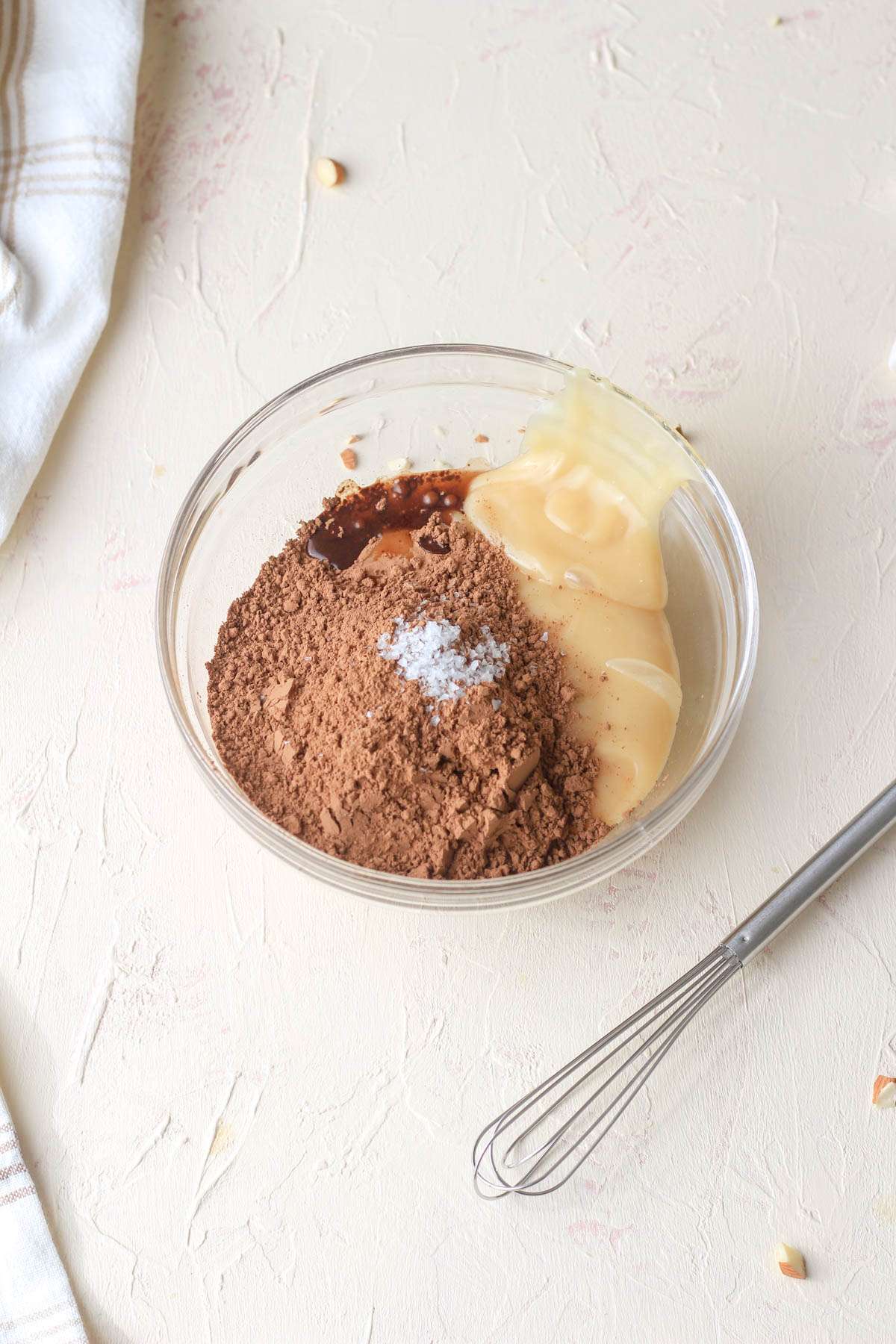 A small glass bowl with a whisk to the right and the ingredients for the sweetened condensed oat milk and cocoa powder mixture.