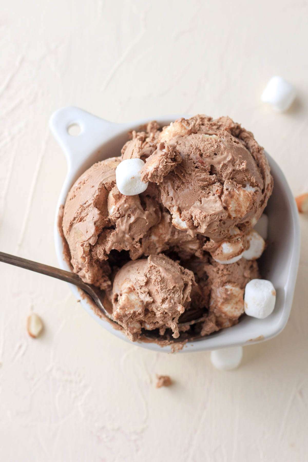 A white bowl of rocky road ice cream on a cream counter with a spoon in the left side of the bowl.