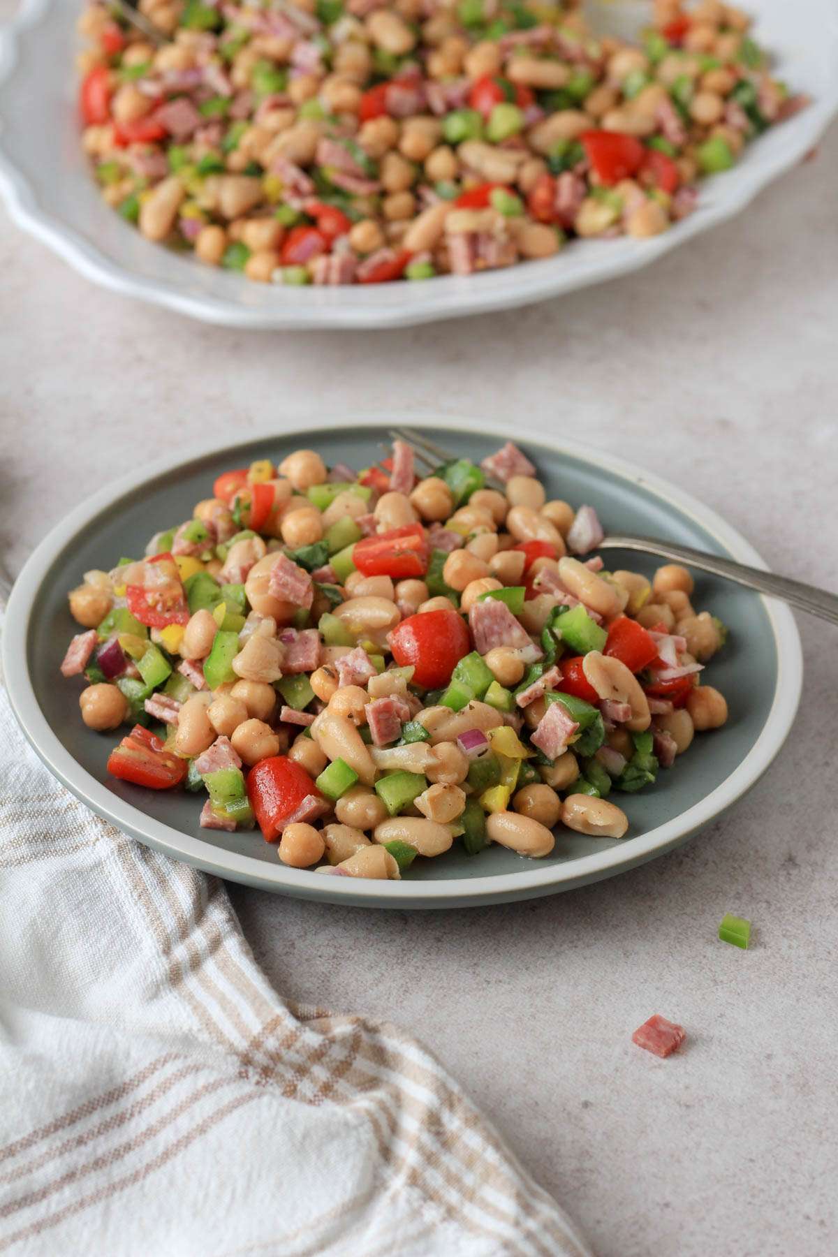 From the side image of a green plate with a serving of Italian grinder bean salad with a silver fork in front of a white platter of bean salad.